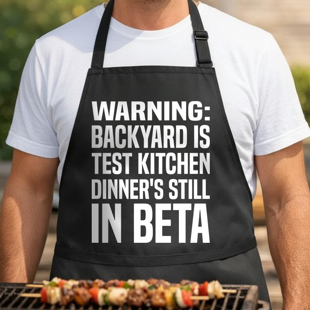Backyard Test Kitchen black BBQ apron, torso close-up: Black apron with large white block letters reading 'Warning Backyard Is Test Kitchen Dinner's Still In Beta' in bold typography.