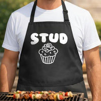 Stud Muffin black BBQ apron, torso close-up: Black apron with white bold text 'STUD' above a detailed line drawing of a cupcake with a rose on top.