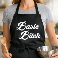 Basic Bitch black BBQ apron, woman torso close-up: Black apron with large white bold script text reading Basic Bitch on front, simple and minimalist design