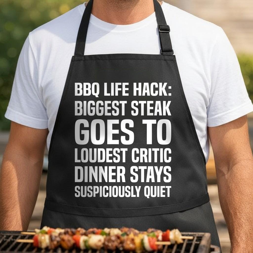 Steak Silence Hack black BBQ apron, torso close-up: Black apron with large white bold text reading a humorous BBQ life hack about giving the biggest steak to the loudest critic for quiet dinner