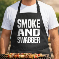 Smoke & Swagger black BBQ apron, torso close-up: Black apron with large white block letters spelling Smoke and Swagger in three stacked lines, bold minimalist style.