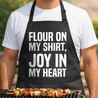 Flour on My Shirt, Joy in My Heart black BBQ apron, torso close-up: Black apron with large white bold text reading Flour On My Shirt, Joy In My Heart in three stacked lines, simple typography design.