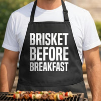 Brisket Before Breakfast black BBQ apron, torso close-up: Black apron with large white bold letters reading Brisket Before Breakfast in three lines, simple typography design