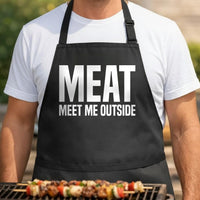 Meat Me Outside black BBQ apron, torso close-up: Black apron with large white text saying Meat Me Outside in bold capital letters, simple typography design.