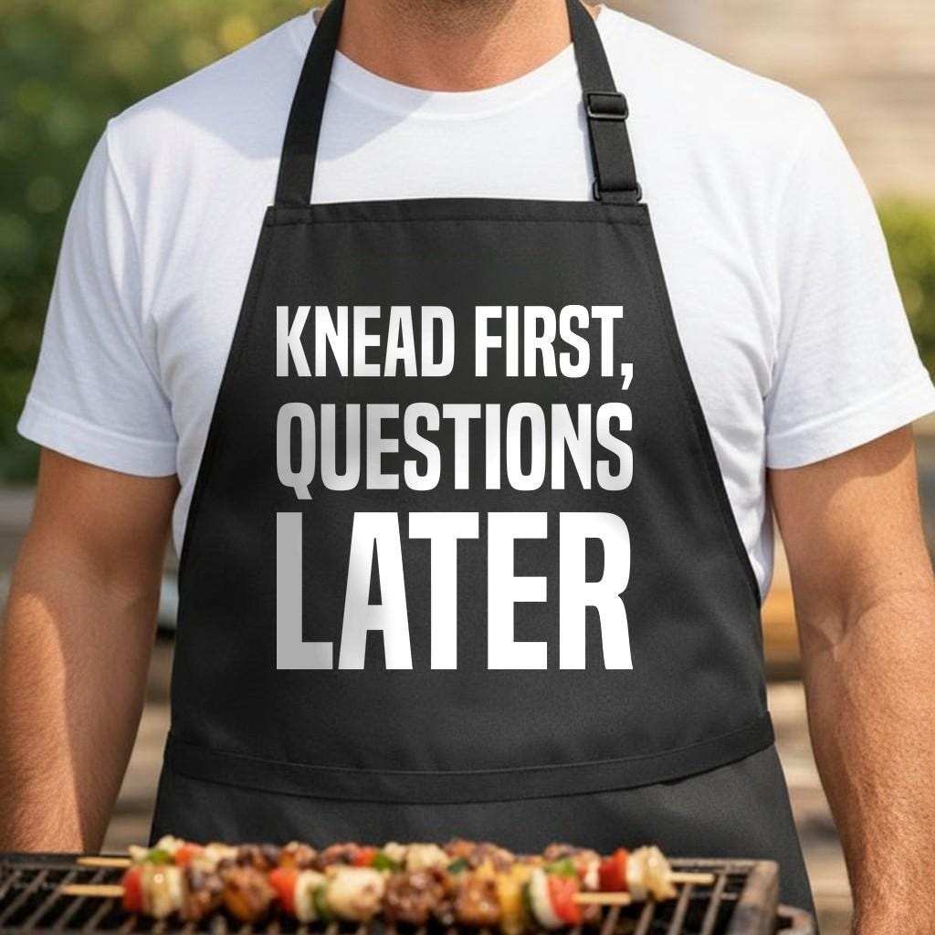 Knead First, Questions Later black BBQ apron, torso close-up: Black apron with large white text reading Knead First, Questions Later in bold typography on a plain background