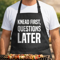 Knead First, Questions Later black BBQ apron, torso close-up: Black apron with large white text reading Knead First, Questions Later in bold typography on a plain background