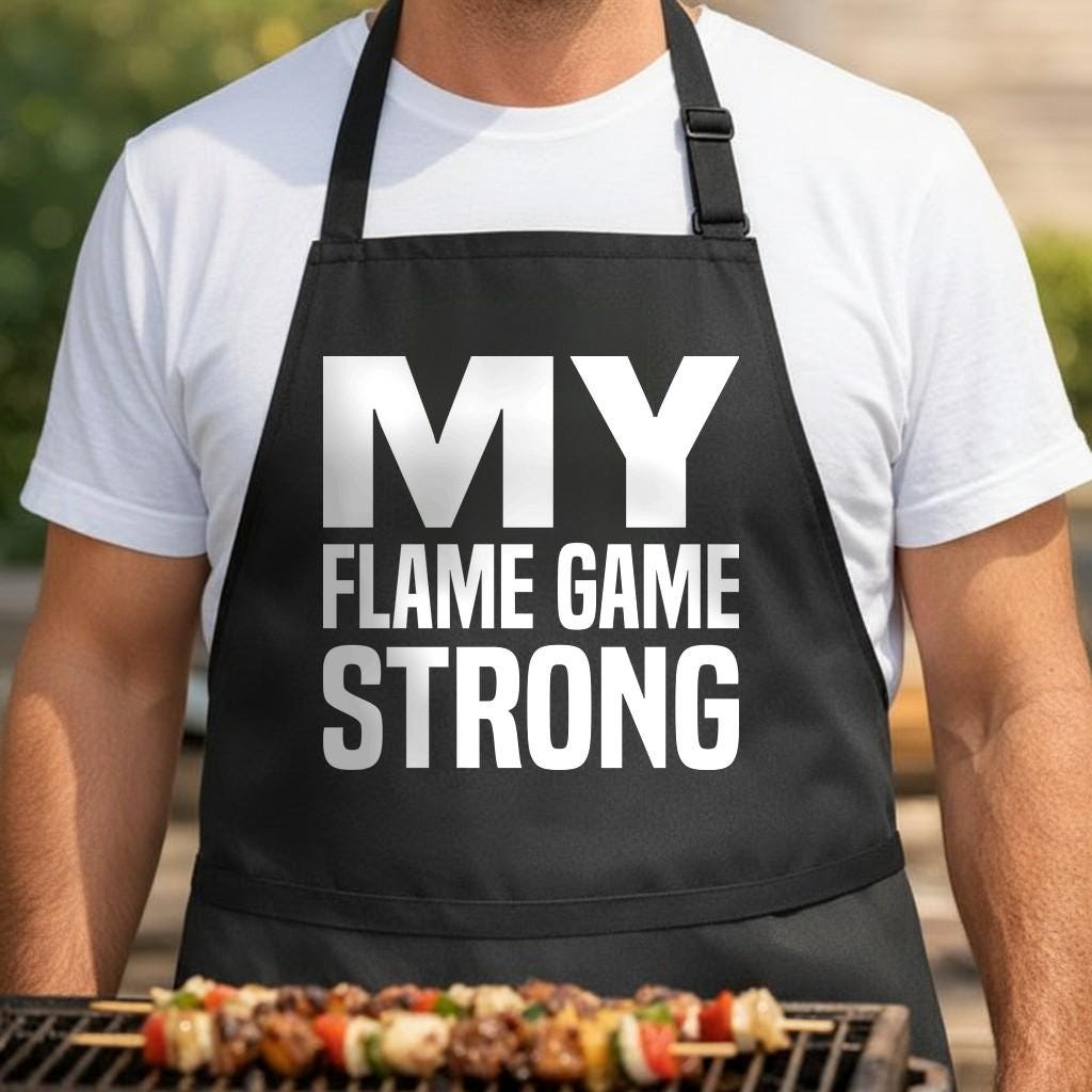 Flame Game Strong black BBQ apron, torso close-up: Black apron with large white text reading My Flame Game Strong in bold capital letters on front