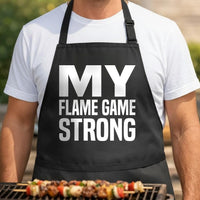 Flame Game Strong black BBQ apron, torso close-up: Black apron with large white text reading My Flame Game Strong in bold capital letters on front