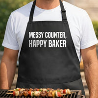 Messy Counter, Happy Baker black BBQ apron, torso close-up: Black apron with large white bold text reading 'Messy Counter, Happy Baker' in all capital letters, simple typographic design.