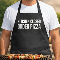 Kitchen Closed, Order Pizza black BBQ apron, torso close-up: Black apron with large white bold text saying Kitchen Closed Order Pizza in a simple, modern style