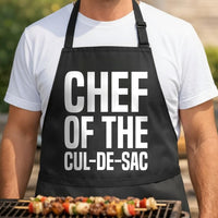 Chef of the Cul-de-Sac black BBQ apron, torso close-up: Black BBQ apron with large white bold text reading 'Chef of the Cul-de-Sac' in a clean, modern font.
