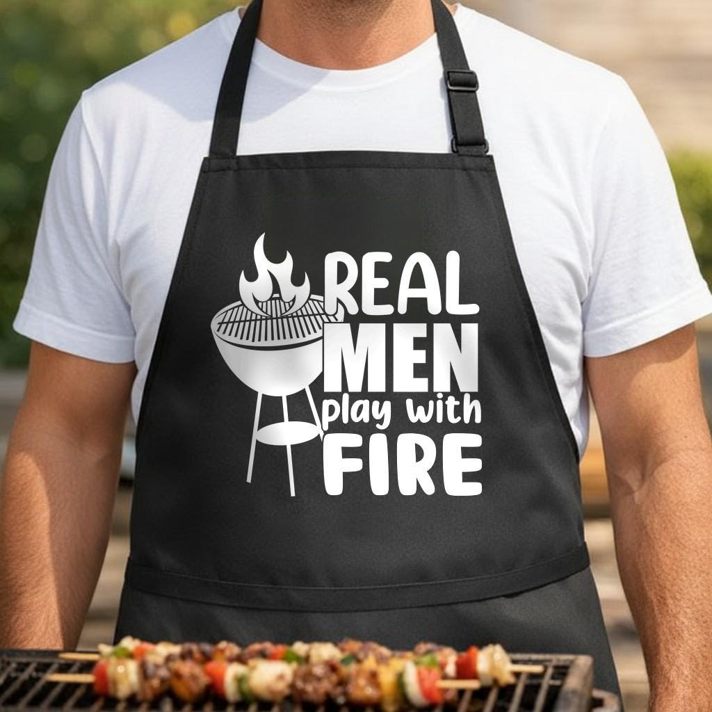 Real Men Play with Fire black BBQ apron, torso close-up: Black apron with white graphic of a charcoal grill with flames and text that reads Real Men Play With Fire in bold letters.