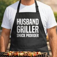 Husband, Griller, Snack Provider black BBQ apron, torso close-up: Black apron with large white bold text saying Husband Griller Snack Provider in three lines on front
