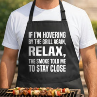 Smoke Told Me black BBQ apron, torso close-up: Black apron with large white text saying, If I'm Hovering By The Grill Again, Relax, The Smoke Told Me To Stay Close.