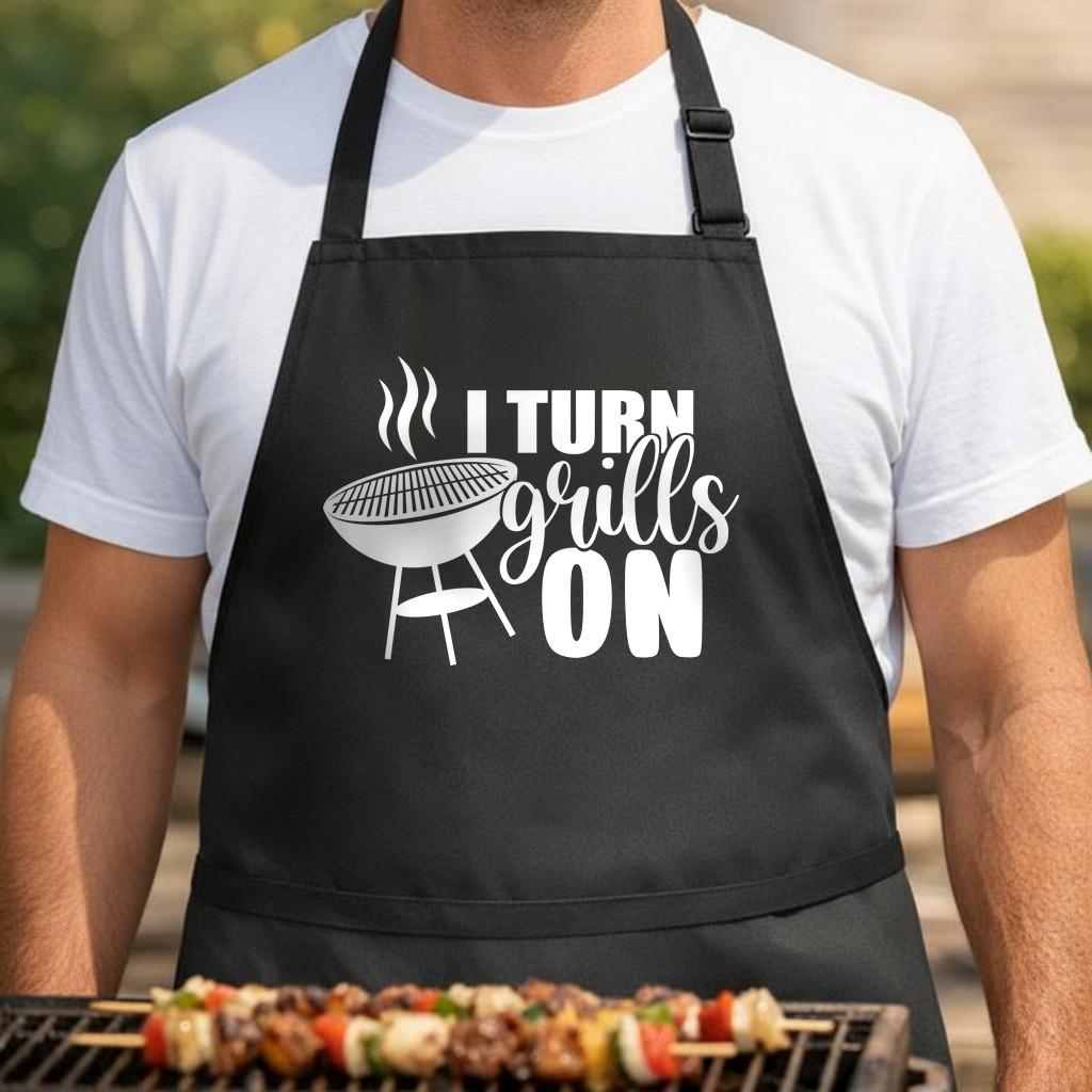 I Turn Grills On black BBQ apron, torso close-up: Black apron with white design showing a steaming charcoal grill and the phrase ‘I Turn Grills On’ in bold and script fonts.