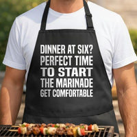 Marinade Time Math black BBQ apron, torso close-up: Black apron with large white block text reading a funny grilling message about starting marinade at dinner time, humorous cooking theme.