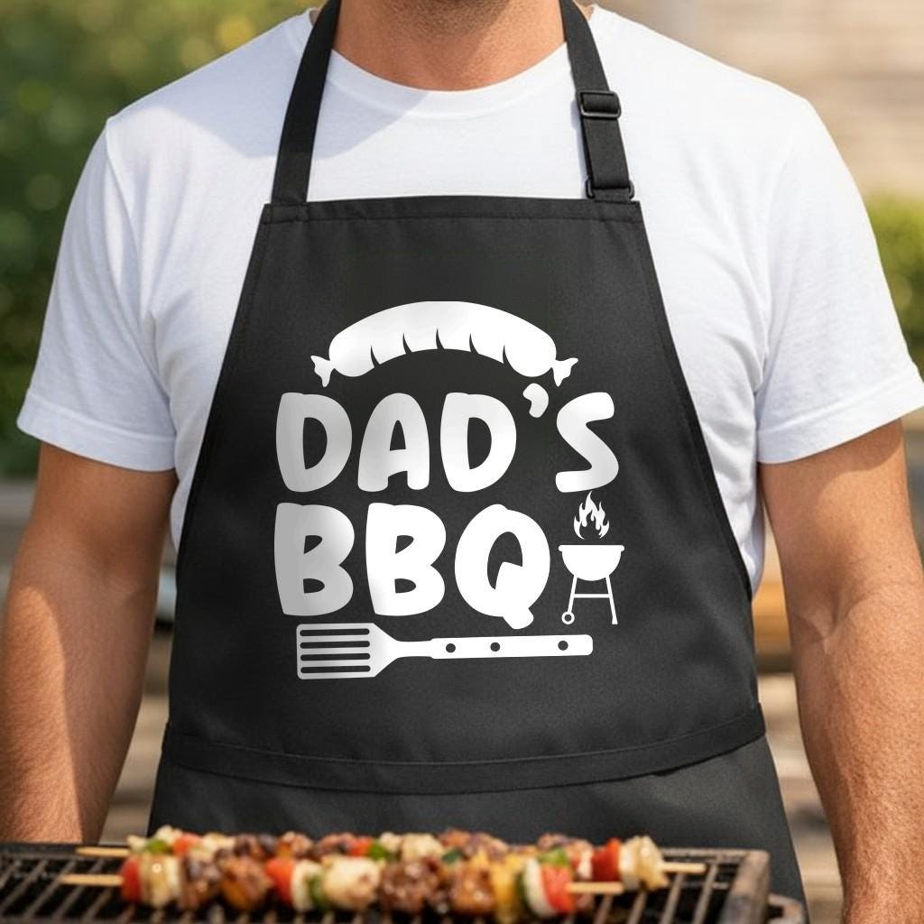 Dad's BBQ black BBQ apron, torso close-up: White graphic on black apron showing a sausage above the words Dad's BBQ with a flaming grill and spatula below in a bold style