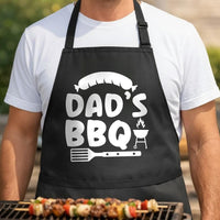 Dad's BBQ black BBQ apron, torso close-up: White graphic on black apron showing a sausage above the words Dad's BBQ with a flaming grill and spatula below in a bold style