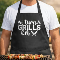 I Turn Grills On black BBQ apron, torso close-up: Black apron with white text saying 'I turn grills on' with flame icons and crossed knives graphic in bold typography