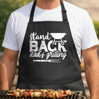 Stand Back Dad's Grilling black BBQ apron, torso close-up: Black apron with white text saying Stand Back Dad's Grilling featuring a kettle grill and barbecue fork graphic in simple vector style.