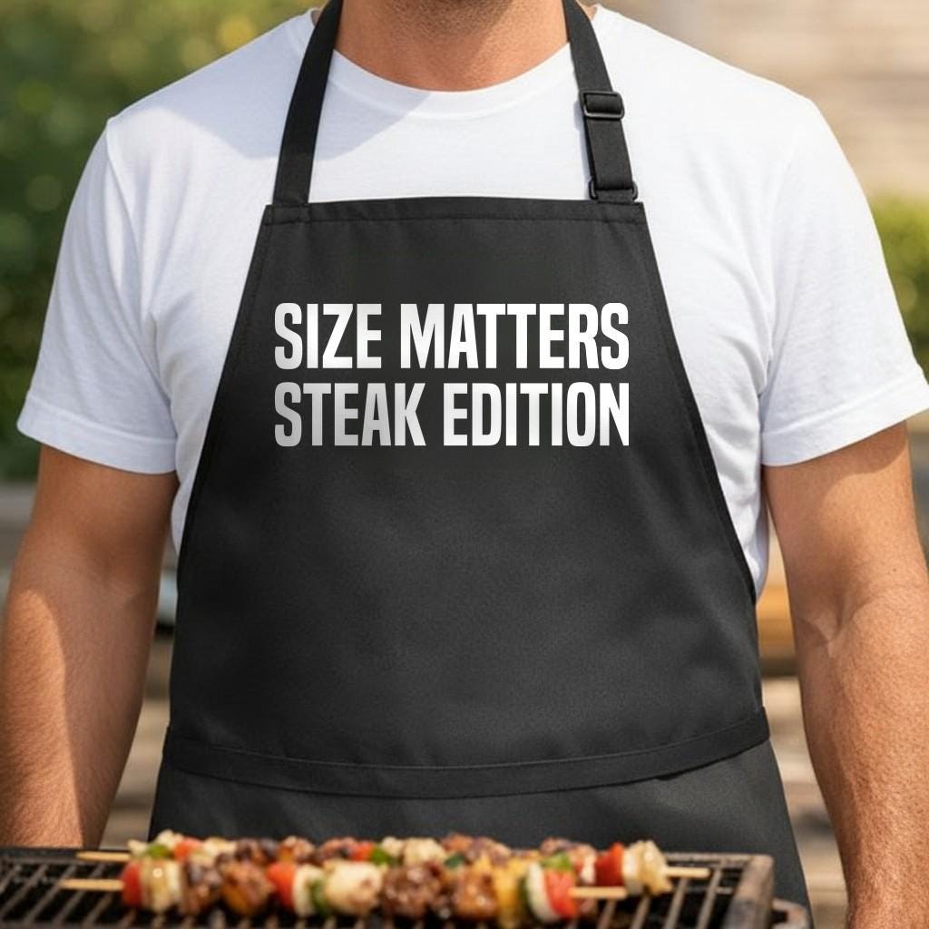 Size Matters: Steak Edition black BBQ apron, torso close-up: Black apron with bold white text saying Size Matters Steak Edition in all capital letters on front