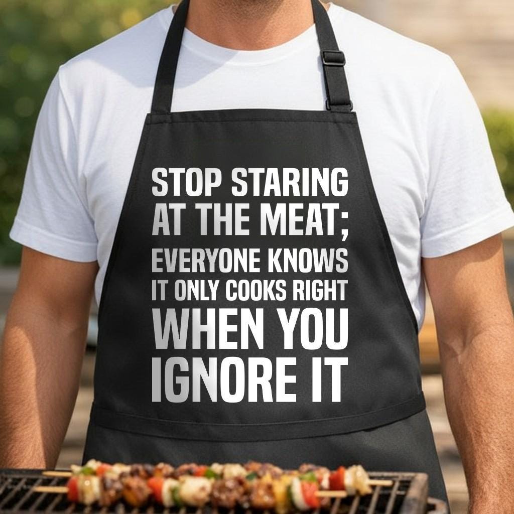Shy Meat Theory black BBQ apron, torso close-up: Black apron with large white bold text reading stop staring at the meat everyone knows it only cooks right when you ignore it