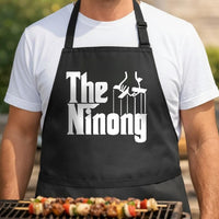 The Ninong black BBQ apron, torso close-up: Black apron with white text 'The Ninong' and a hand controlling puppet strings in bold graphic design.