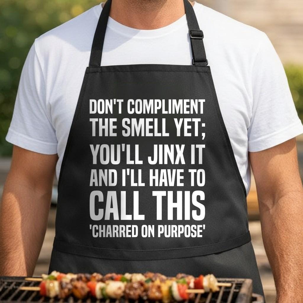 Don't Jinx Dinner black BBQ apron, torso close-up: Black apron with large white bold text saying do not compliment the smell yet you'll jinx it and I'll have to call this charred on purpose