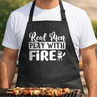 Real Men Play with Fire black BBQ apron, torso close-up: Black apron with white bold and script text reading Real Men Play With Fire and a small grill with flames icon